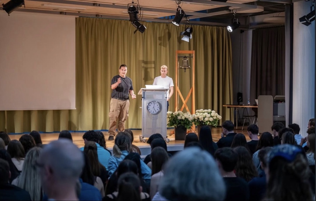 Nikola Knežević hosting the Uselüti ceremony on stage at Kantonsschule Zofingen, standing next to a wooden stand with the traditional farewell bell and a lectern, addressing a full auditorium of students and parents.